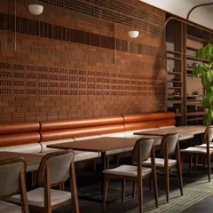 This seating area features a long, comfortable booth against a textured terracotta brick wall. The combination of warm leather-toned upholstery and natural wood chairs creates a cozy and inviting zone for guests to enjoy their meal.