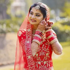 This outdoor bridal portrait captures a quiet, candid moment. The bride is adjusting her earring, a simple gesture that feels natural and elegant. The soft, natural light beautifully illuminates her red embroidered suit and the intricate henna on her hands.