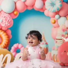 The pure joy of a first birthday cakesmash. This little girl's happy, messy smile is captured against a vibrant candy-themed backdrop with pink and blue balloons, showing a fully styled milestone session.