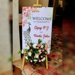 A beautifully designed welcome sign for an engagement, featuring an elegant peacock illustration. The sign is complemented by a lovely arrangement of pink and white flowers, adding a graceful touch.