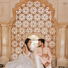 An intimate moment framed by a stunningly intricate marble lattice (jaali) in a Jaipur palace. The soft light filtering through creates a dreamy and romantic atmosphere for this pre-wedding portrait.