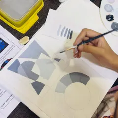 Here, a student is working on a value study exercise. This involves painting geometric shapes with a range of tones from black to white, which is essential for understanding depth and form.
