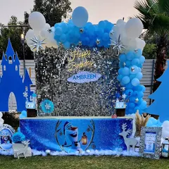 A magical Frozen-themed birthday party setup. The centerpiece is a shimmering silver sequin backdrop, framed by a blue and white balloon garland, with cutouts of Elsa's castle and other winter elements to create an enchanting wonderland.
