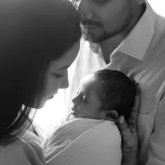 A new family, bathed in soft light. This black and white portrait emphasizes the tender, protective embrace of the parents around their sleeping newborn.