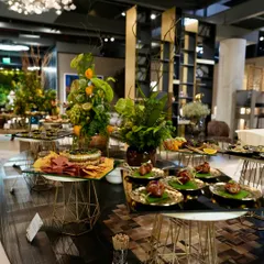 A wide shot of our catering setup for a luxury brand event. Multiple stations with geometric stands display a variety of appetizers and grazing boards, creating an immersive culinary landscape.