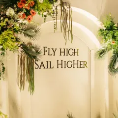 This custom arched backdrop for a joint 50th and 80th birthday features a personal quote, "Fly High Sail Higher," framed by lush tropical greenery and florals, making it a meaningful centerpiece.