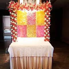 A vibrant floral mandap for a home Ganpati celebration. The backdrop is made of colorful yellow and pink flower panels, framed by artificial rose garlands.