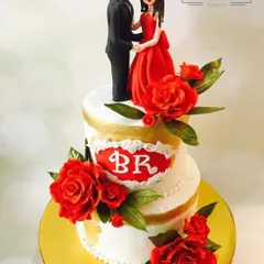 A love story told in sugar. This elegant two-tiered wedding cake features a custom couple topper in a red dress and suit, surrounded by beautiful red sugar roses, perfect for a romantic celebration.