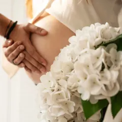 A close-up shot focusing on the hands of the couple gently holding the baby bump, with a bouquet of white flowers adding a touch of elegance and softness.