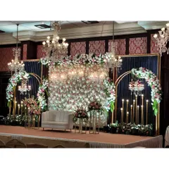 This elegant reception stage features a full wall of white and blush florals, framed by modern gold arches and accented with hanging crystal chandeliers. The design combines classic romance with contemporary style, providing a luxurious backdrop for the couple.