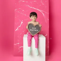 A fun and modern "Big Brother" announcement. A smiling boy sits against a vibrant pink backdrop holding a heart-shaped chalkboard, excited for his new role.