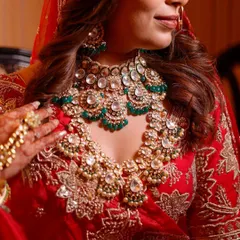 A close up shot focusing on the bride's magnificent jewelry. The layered Kundan necklace with green stones perfectly complements her red embroidered lehenga, showcasing royal elegance.