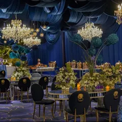 A wide view of a Victorian-inspired blue cocktail event. The space is defined by deep blue fabric draping, opulent crystal chandeliers, and dramatic floral centerpieces, creating a regal and luxurious atmosphere.