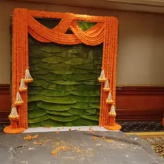 I create authentic backdrops that blend natural elements with vibrant flowers. This piece features a wall of fresh banana leaves framed by draped orange marigold garlands and hanging jasmine tassels, perfect for a traditional pooja or event.