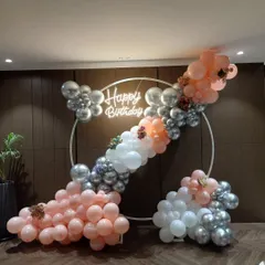 An elegant circular backdrop for a birthday celebration. The white metal ring is decorated with a garland of pastel pink, white, and silver chrome balloons, with a "Happy Birthday" neon sign in the center.