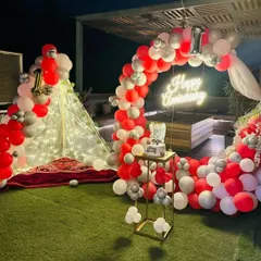 A first-anniversary party on a terrace with an artificial lawn. The setup features a circular balloon arch in red and white, a neon sign, and a cozy, lit-up teepee tent.
