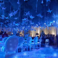 A wide view of the 'VManTakesUdaan' themed event. The entire ceiling is a sea of blue, with hanging origami birds and crystals, creating a surreal, underwater-like atmosphere for the guests.