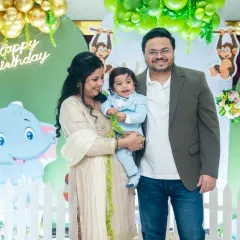 A happy family portrait at a jungle-themed first birthday party, with the parents and birthday boy posing together in front of a vibrant green and gold balloon backdrop.