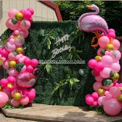 An outdoor flamingo-themed party with a green hedge wall backdrop, a neon sign, and bright pink balloon garlands.