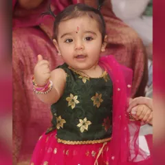 A close-up portrait of a baby girl in a beautiful traditional lehenga, giving a little thumbs-up.
