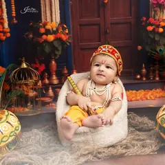 A classic Little Krishna pose. The baby sits in a miniature chair, holding a flute, surrounded by a vibrant, festive setup complete with peacock feathers and decorated pots.