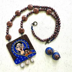 This piece, named 'Mamta', is a celebration of a mother's love. The hand-painted pendant shows a tender moment between mother and child, paired with matching wooden jhumkas and finished with delicate shells that add a soft, natural touch.
