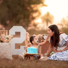 An outdoor cake smash for a second birthday. A mother helps her son taste the cake in a beautiful, natural setting during golden hour.