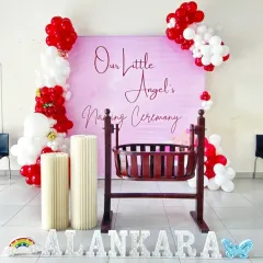 A sweet and simple naming ceremony for a baby girl. The pink backdrop is framed by red and white balloons, with a traditional wooden cradle as the centerpiece and marquee letters spelling the name.