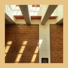 Beams of sunlight stream in from high clerestory windows, creating strong diagonal lines across an exposed brick wall. This technique allows for natural illumination while maintaining privacy in the spaces below.