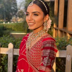 A breathtaking bride captured in the soft, natural light of her outdoor wedding. The makeup is designed to be luminous and fresh, complementing her red lehenga and floral bun.