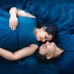 An artistic overhead shot of an expecting couple lying together, enveloped in the rich fabric of a deep blue gown. This pose captures a quiet, intimate moment of connection.