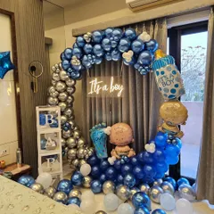 A stylish and modern "It's a Boy" welcome home decoration. This setup features a half-moon balloon arch in metallic blue and silver, a neon sign, and cute props like baby foil balloons and transparent 'BABY' blocks.