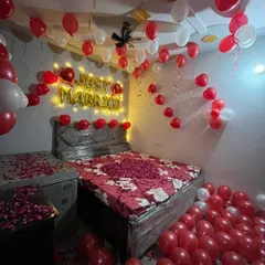 A classic red and white themed 'Just Married' room decoration. This setup includes 'Just Married' foil balloons with warm backlighting, a bed covered in a rose petal heart, and the floor filled with balloons.