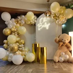 This 'Oh Baby' shower setup is a popular choice, featuring a soft and elegant color palette of white, cream, and chrome gold balloons. The arrangement is built around a white arched backdrop with a neon sign and includes a large teddy bear prop and golden pedestals for a beautiful display.