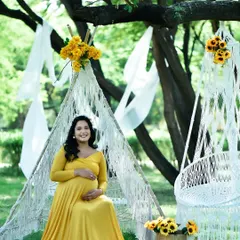 A bright and cheerful outdoor pregnancy photo. The expectant mother is wearing a lovely yellow gown, seated in a bohemian-style setup with a macrame teepee and hanging chair.