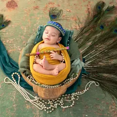 A sleeping newborn styled as Krishna, holding a flute and nestled in a basket with a beautiful peacock feather arrangement.