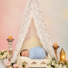 A baby sleeps peacefully in a miniature bed under a delicate lace tent, surrounded by candles and flowers. This setup is perfect for a romantic and whimsical newborn photoshoot.
