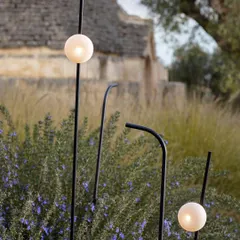 The Firefly outdoor lights by Karman, resembling glowing spheres on thin black stems, are nestled among lavender bushes. This image shows how these lights can be used to create a magical, firefly-like effect in a garden at dusk.