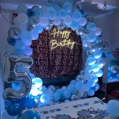 A classic circular balloon backdrop for a 5th birthday. The frame is covered in blue and silver balloons and features a 'Happy Birthday' neon sign, making it a simple and elegant photo corner for a home party.