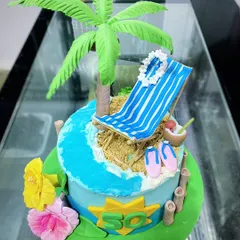A relaxing beach-themed cake for a 50th birthday. This design features a fondant beach chair, palm tree, and the ocean shore, all completely edible.