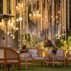 A cozy lounge area set up for a wedding ceremony, under a canopy of cascading mogra and lit by the warm glow of chandeliers. The natural wood furniture adds a touch of rustic elegance to the traditional setting.