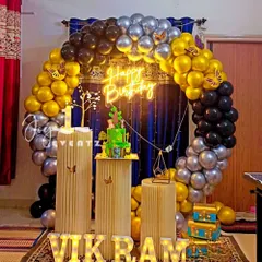 I created this elegant balloon ring decoration for Vikram's first birthday. The arch of gold, black, and silver balloons is set against a dark curtain, with fluted plinths for the cake and light-up marquee letters.