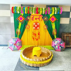 This is a classic Haldi ceremony setup. It features a bright yellow and green backdrop, a traditional brass urli for the ritual, and decorative umbrellas, creating a perfect, joyful setting for your function.