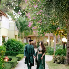 A couple in matching deep green traditional wear walks hand-in-hand through the lush gardens of the Novotel in Goa.