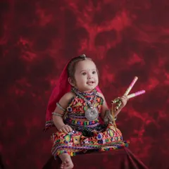This little one is ready for Navratri. Dressed in a colorful traditional outfit and holding dandiya sticks, she is the star of her own festive toddler shoot against a rich, textured red backdrop.