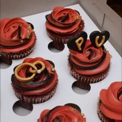 A detailed view of the engagement-themed cupcakes. This shot highlights the handcrafted fondant rings and the heart toppers with the couple's initials, set against a rich, red buttercream rose swirl.