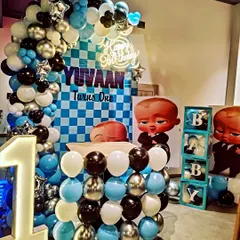 A cool Boss Baby themed first birthday setup. The decoration uses a blue, black, and silver color scheme with a checkered backdrop, a large balloon arch, 'BABY' blocks, and a light-up number '1'.