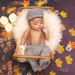 This autumn-themed portrait features a baby boy dressed in a vintage-style flat cap and suspenders. He is resting in a basket surrounded by fall leaves, pinecones, and soft candlelight.
