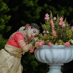 A serene bridal portrait taken outdoors. The bride, dressed in a traditional silk saree, rests against a floral arrangement, creating a peaceful and artistic image that highlights her grace and beauty.