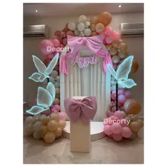 A stunning butterfly-themed backdrop for Ayzal's first birthday. The setup features a large pink bow, glowing neon butterflies, and a cascade of pink and peach balloons.